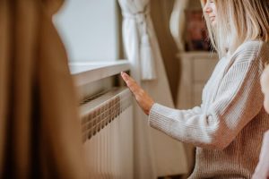 Young blond woman in long winter beige sweater is posing at home near the radiator.