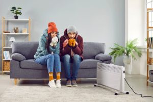 A young man and woman sitting on sofa in the living room in winter outerwear and in hats at home warming their hands from a space heater. Heating problems and broken furnace concept.