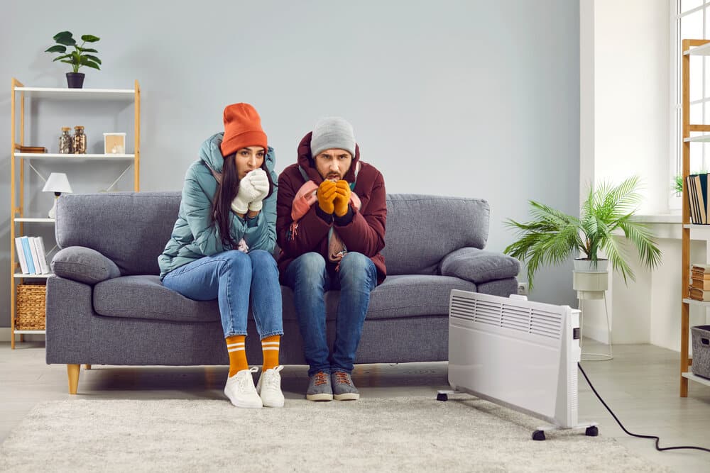 A young man and woman sitting on sofa in the living room in winter outerwear and in hats at home warming their hands from a space heater. Heating problems and broken furnace concept.