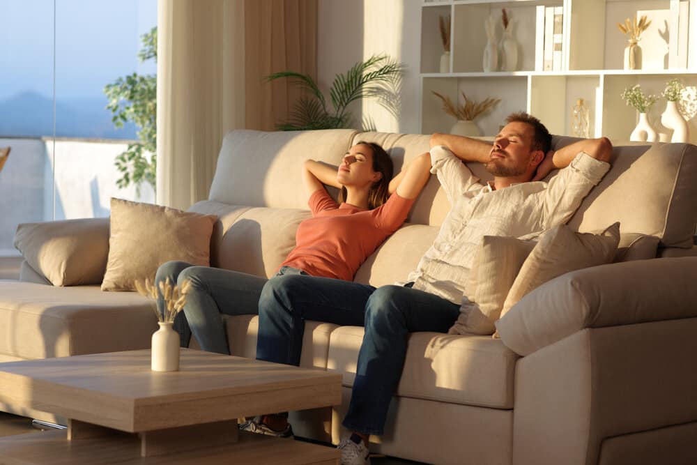 Happy couple relaxing sitting on a couch with arms on head at sunset at home